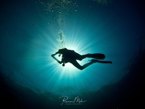Ein Taucher schwebt als Silhouette vor der Sonne im blauen Wasser und erzeugt dabei Luftblasen. Die dramatische Gegenlichtaufnahme zeigt die Sonnenstrahlen, die durch das Wasser brechen und eine mystische Atmosphäre schaffen.
