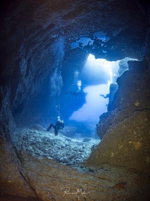Mehrere Taucher schwimmen durch den Ausgang der großen Höhle hinter dem berühmten Blue Hole auf Gozo ins Freie. Luftblasen haben sich an der Höhlendecke gesammelt und bilden eine spiegelnde Oberfläche