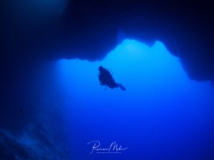 Ein Taucher schwebt als Silhouette vor dem spektakulären blauen Licht einer Höhlenöffnung. Die dramatische Beleuchtung erzeugt einen eindrucksvollen Kontrast zwischen der dunklen Höhle und dem strahlend blauen Wasser im Hintergrund.
