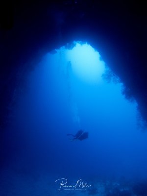 Blick aus einer Unterwasserhöhle heraus ins offene Wasser, wobei das einfallende Sonnenlicht ein spektakuläres blaues Lichtspiel erzeugt. Im mittleren Bereich ist die Silhouette eines Tauchers zu erkennen, der durch das tiefblaue Wasser gleitet.