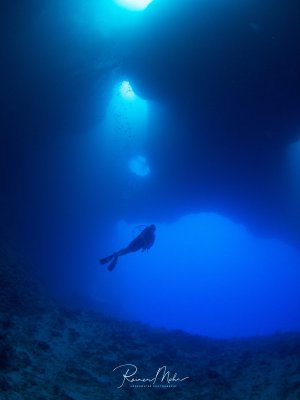 Ein Taucher schwebt als dunkle Silhouette im tiefblauen Wasser einer Unterwasserhöhle und blickt zu hellen Lichtstrahlen auf, die von der Wasseroberfläche durchdringen. Die atmosphärische Szene zeigt den Kontrast zwischen der dunklen Tiefe und dem hellen Sonnenlicht von oben.