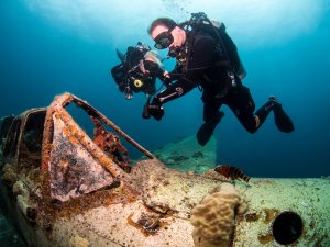 Zwei Taucher mit Kameras erkunden ein stark bewachsenes Wrack am Meeresboden. Das Flugzeugwrack ist mit Korallen und anderen Meeresorganismen überzogen und bietet Lebensraum für verschiedene Rifftiere, die zwischen den rostigen Strukturen zu erkennen sind.