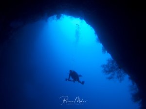 Ein Taucher schwimmt durch eine beeindruckende Unterwasserhöhle, wobei das intensive blaue Licht vom Höhlenausgang eine dramatische Silhouette erzeugt. Die Szene zeigt die majestätische Dimension der Unterwasserlandschaft mit ihren natürlichen Felsformationen.