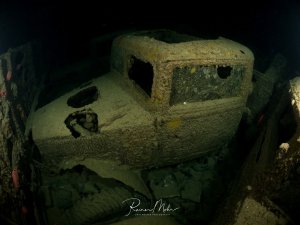 Ein altes Fahrzeug im Inneren des Wracks der SS Thistlegorm, dick mit Sediment und Korrosion überzogen. Die Kabine und die Räder sind noch gut erkennbar, die Atmosphäre ist geheimnisvoll.