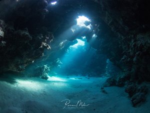 Ein faszinierender Blick aus einer Unterwasserhöhle heraus, bei dem dramatische Lichtstrahlen durch die Öffnungen fallen und den sandigen Höhlenboden sowie die zerklüfteten Felswände in ein mystisches blaues Licht tauchen. Die natürliche Kathedrale aus Stein schafft eine beeindruckende Atmosphäre mit mehreren Ausgängen zum offenen Wasser.