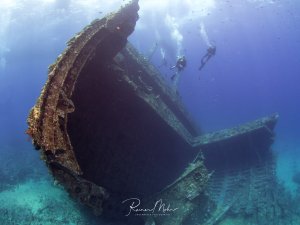 Ein imposantes Schiffswrack liegt auf sandigem Meeresgrund, dessen Bug dramatisch zur Wasseroberfläche zeigt. Zwei Taucher erkunden das mit Korallen und Meeresorganismen bewachsene Wrack, das zu einem künstlichen Riff geworden ist.