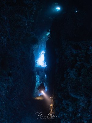 Ein Taucher schwebt durch einen engen Spalt in einer Unterwasserhöhle. Das blaue Licht vom Eingang und die Taucherlampe beleuchten die bewachsenen Felswände.