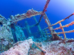 Ein versunkenes Schiffswrack, das vollständig von bunten Korallen und Meeresorganismen bewachsen ist. Die Metallstrukturen des ehemaligen Schiffs sind noch deutlich erkennbar, während kleine bunte Riffische um das künstliche Riff schwimmen.