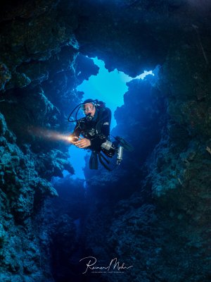 Ein Taucher mit Unterwasserkamera schwebt im Canyon des Thomas-Riffs im Roten Meer. Das blaue Licht von außen rahmt die Silhouette des Tauchers mit seiner Lampe ein.