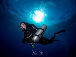 Ein Taucher schwebt im tiefblauen Wasser mit seitlicher Stage-Flasche, während Sonnenstrahlen von oben durch die Wasseroberfläche dringen und eine dramatische Lichtstimmung erzeugen. Die Aufnahme zeigt die klassische Silhouette eines Tauchers gegen das helle Oberflächenlicht.