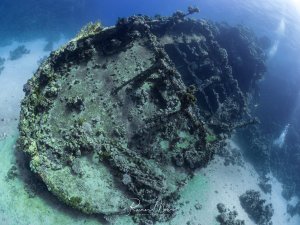Weitwinkelaufnahme eines großen, mit Korallen bewachsenen Wracks am Meeresgrund des Roten Meeres. Die Struktur des Schiffskörpers ist unter dem Bewuchs noch erkennbar, ein Taucher schwebt daneben.