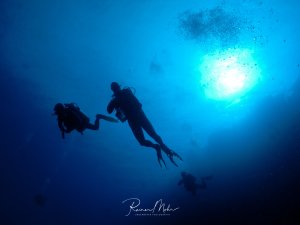 Drei Taucher schweben im tiefblauen Wasser während eines Sicherheitsstopps oder Aufstiegs zur Oberfläche. Die Szene wird dramatisch von den Sonnenstrahlen beleuchtet, die von oben durch das Wasser dringen und eine mystische Atmosphäre schaffen.