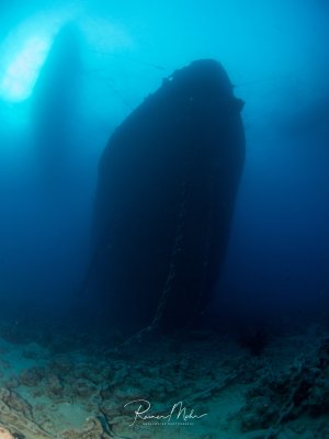 Ein imposantes Unterwasserwrack ragt als dunkle Silhouette gegen das blaue Meerwasser auf. Die dramatische Aufnahme zeigt den Bug des versunkene Schiffs vom Meeresgrund aus betrachtet, mit Sonnenstrahlen die durch das Wasser dringen und eine mystische Atmosphäre schaffen.