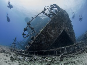 Das Wrack der Giannis-D am Riff Abu Nuhas im Roten Meer liegt geneigt auf dem Meeresboden, während mehrere Taucher das imposante Wrack erkunden. Das Schiff ist bereits stark von Meeresorganismen bewachsen und zeigt deutliche Korrosionsspuren, die Aufbauten und Reling sind noch gut erkennbar.