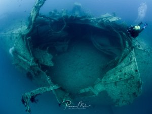 Ein Taucher erkundet die imposante Bombeneinschlagstelle des Wrack der Rosalie Moller, die aufrecht auf dem Meeresboden steht. Die massive Struktur mit ihren Aufbauten ist bereits von Meeresorganismen besiedelt.