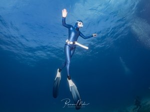 Freitaucherin in blauem Anzug steigt elegant im Blauwasser auf, eine Hand erhoben. Im Hintergrund ein weiterer Taucher.