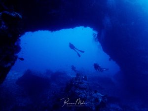 Taucher erkunden den Torbogen (Arch) am Elphinstone Riff, der von intensivem blauem Licht durchflutet wird. Die Silhouetten der Taucher heben sich dramatisch gegen das helle Blau des Höhlenausgangs ab, während die Felswände im Vordergrund die natürliche Architektur der Unterwasserlandschaft zeigen.