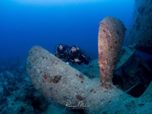 Zwei Taucher erkunden die Schiffsschraube eines großes Schiffswracks in tiefem blauen Wasser. Die Metallteile des Wracks sind stark mit Meeresorganismen bewachsen und zeigen deutliche Korrosionsspuren, was auf das Alter des gesunkenen Schiffes hinweist.