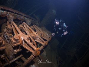 Ein Taucher erkundet ein historisches Schiffswrack und betrachtet den rostigen Schiffsdiesel des Wracks. Die Metallteile sind stark korrodiert und mit marinen Aufwuchs bedeckt, was auf das hohe Alter des Wracks hindeutet.